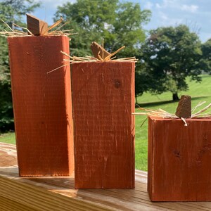 May include: Three wooden pumpkin decorations in varying sizes. Each pumpkin is a rectangular block painted in a warm orange hue, with a small wooden stem and straw accents on top. The pumpkins are arranged on a wooden railing.