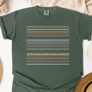 May include: A moss green t-shirt with a geometric pattern of horizontal lines in various colors, including peach, tan, and gray. The word "MOSS" is printed on a beige circle. A straw hat is visible in the bottom right corner.