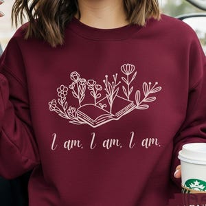 May include: A garnet-colored sweatshirt features a white floral design with an open book and the text "I am, I am, I am." The sweatshirt is a crew neck style and has long sleeves. A coffee cup with a brown sleeve is held in the hand.