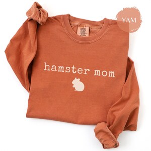May include: A rust-colored long-sleeve shirt with the words "hamster mom" printed in white, along with a white hamster graphic. The shirt has a crew neck and rolled-up sleeves. The shirt is made of a soft material.