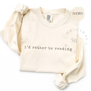 May include: Ivory-colored long-sleeve shirt with the text "i'd rather be reading" in a simple font. The shirt has a crew neck and a tag that says "Comfort Colors". The sleeves are long and the shirt is folded.