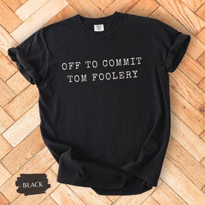 May include: Black t-shirt with the text "OFF TO COMMIT TOM FOOLERY" in white. The shirt is displayed on a wooden surface. The word "BLACK" is written on a black rectangle in the bottom left corner.