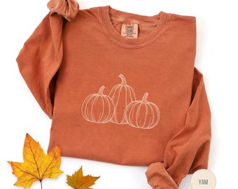 Puede incluir: Una camiseta de manga larga naranja quemado con un contorno blanco de tres calabazas. La camiseta tiene cuello redondo y mangas enrolladas. El color de la camiseta es "Yam" según la etiqueta. Dos hojas de otoño amarillas y naranjas están en la esquina inferior izquierda.