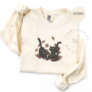 Puede incluir: Camiseta de manga larga color marfil con un gr&aacute;fico de tres gatos negros jugando entre hojas de oto&ntilde;o. Los gatos tienen ojos amarillos. La camiseta tiene cuello redondo y la marca "Comfort Colors" es visible.