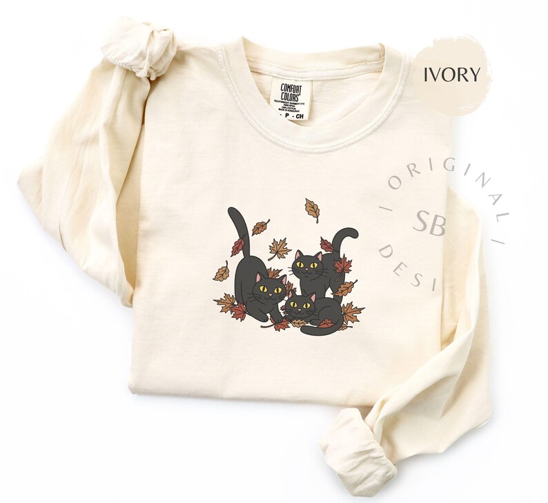 Puede incluir: Camiseta de manga larga color marfil con un gr&aacute;fico de tres gatos negros jugando entre hojas de oto&ntilde;o. Los gatos tienen ojos amarillos. La camiseta tiene cuello redondo y la marca "Comfort Colors" es visible.