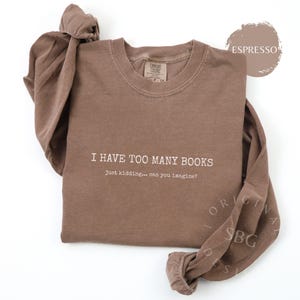 May include: An espresso brown long-sleeve crewneck sweatshirt featuring the white text I HAVE TOO MANY BOOKS and Just kidding... can you imagine? printed on the front. The garment has a ribbed collar and long sleeves with gathered cuffs.