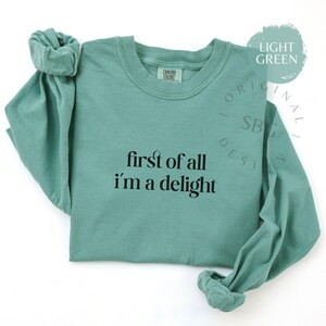 May include: Light green long-sleeve shirt with the text "first of all I'm a delight" in black. The shirt has a crew neck and rolled cuffs. The Comfort Colors tag is visible on the neckline. The shirt is displayed on a white background.