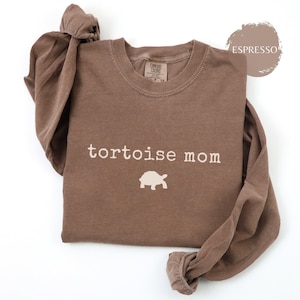 Könnte beinhalten: Espressofarbenes Langarmshirt mit den Worten "tortoise mom" und einer Schildkröten-Grafik in Weiß aufgedruckt. Das Shirt besteht aus einem weichen Material und hat einen Rundhalsausschnitt. Das Shirt ist gefaltet.