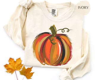 Puede incluir: Sudadera de manga larga color marfil con un diseño de calabaza pintada en tonos naranja, rojo y verde. La sudadera tiene cuello redondo y mangas largas. Dos hojas de otoño amarillas están junto a la sudadera.