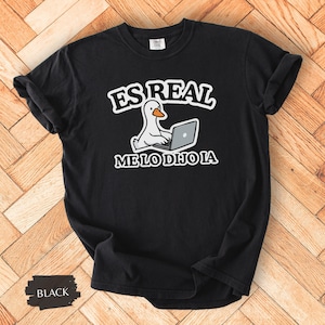 May include: Black t-shirt with a white graphic of a duck using a laptop, and the text "ES REAL ME LO DIJO IA" in white. The shirt has short sleeves and a crew neck.