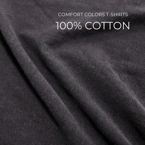 Puede incluir: Primer plano de una tela gris oscuro con textura y pliegues visibles. El texto blanco indica "COMFORT COLORS T-SHIRTS" y "100% COTTON", indicando el material y el tipo de prenda.