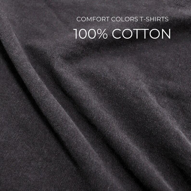 Puede incluir: Primer plano de una tela gris oscuro con textura y pliegues visibles. El texto blanco indica "COMFORT COLORS T-SHIRTS" y "100% COTTON", indicando el material y el tipo de prenda.