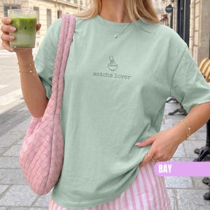 May include: A light green short-sleeve t-shirt featuring a small dark green graphic of a matcha bowl and whisk with the text "matcha lover" printed below it. The shirt is paired with a woven pink shoulder bag and pink and white striped pants.