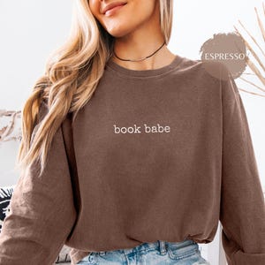 May include: A brown long-sleeve crewneck sweatshirt featuring the text "book babe" embroidered in white on the center chest. The garment is labeled with the color "ESPRESSO" in a circular graphic located in the upper right corner of the image.