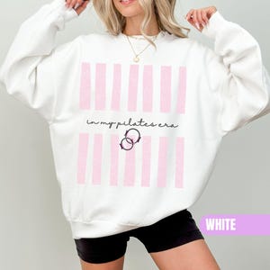 Könnte beinhalten: Ein weißes Langarm-Sweatshirt mit einem Grafikdruck aus vertikalen hellrosa Streifen. Der Text "in my pilates era" ist mittig in schwarzer Schreibschrift aufgedruckt, ergänzt durch eine kleine Abbildung von zwei ineinandergreifenden schwarzen Kreisen.