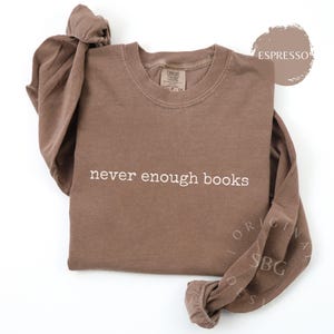 May include: An espresso-colored long-sleeve crewneck sweatshirt featuring the text "never enough books" in white lettering across the chest. The garment has a ribbed neckline and cuffs, with a Comfort Colors brand label visible at the inner collar.