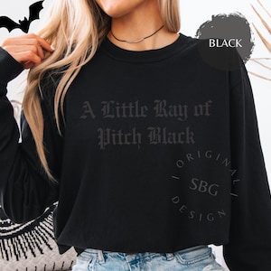 以下が含まれることがあります： ゴシック体で「A Little Ray of Pitch Black」と書かれた黒いスウェットシャツ。黒いコウモリのグラフィックが左上にあります。「BLACK」の文字が右上の黒い円の中にあります。