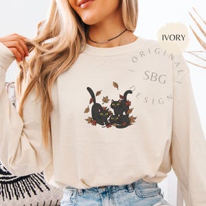 Puede incluir: Camiseta de manga larga color marfil con un gr&aacute;fico de tres gatos negros jugando entre hojas de oto&ntilde;o. Los gatos tienen ojos amarillos. La camiseta tiene el texto "ORIGINAL - SBG DESIGN".