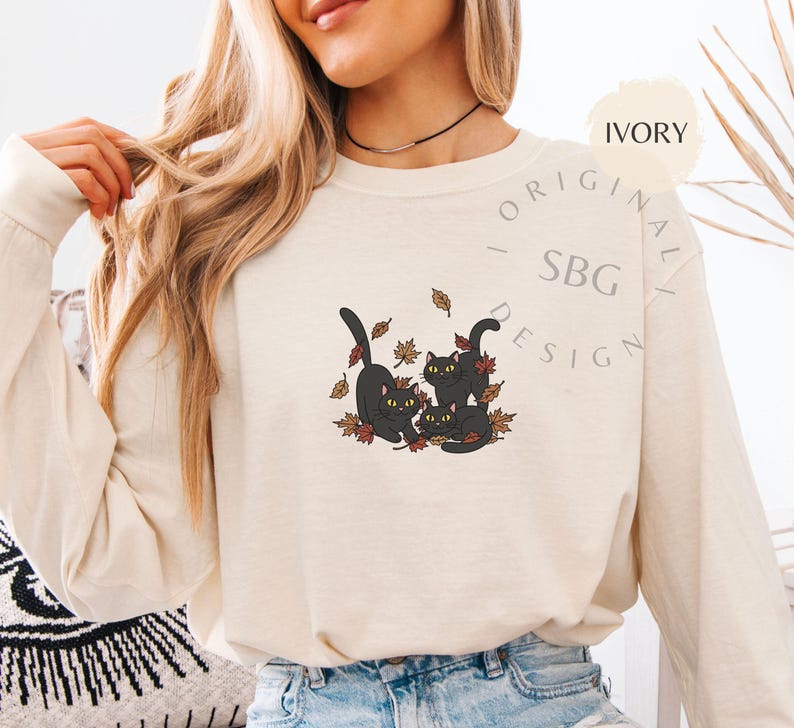 Puede incluir: Camiseta de manga larga color marfil con un gr&aacute;fico de tres gatos negros jugando entre hojas de oto&ntilde;o. Los gatos tienen ojos amarillos. La camiseta tiene el texto "ORIGINAL - SBG DESIGN".