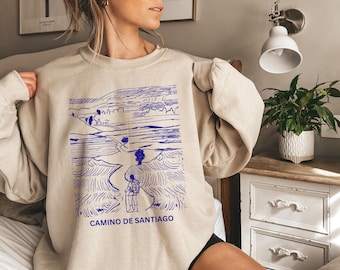 May include: A beige sweatshirt with a blue graphic of a person walking on a path through a landscape. The text "CAMINO DE SANTIAGO" is printed below the graphic.