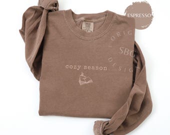 Puede incluir: Camiseta de manga larga color espresso con las palabras "cozy season" y un gráfico de libro. La camiseta tiene cuello redondo y está hecha de un material suave. La marca "ORIGINAL SBG DESIGN" está impresa en la camiseta.