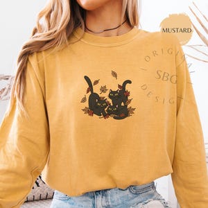 Puede incluir: Sudadera de color mostaza con un gr&aacute;fico de tres gatos negros jugando entre hojas de oto&ntilde;o. El dise&ntilde;o incluye el texto "ORIGINAL SBG DESIGN". La sudadera est&aacute; hecha de un material suave.
