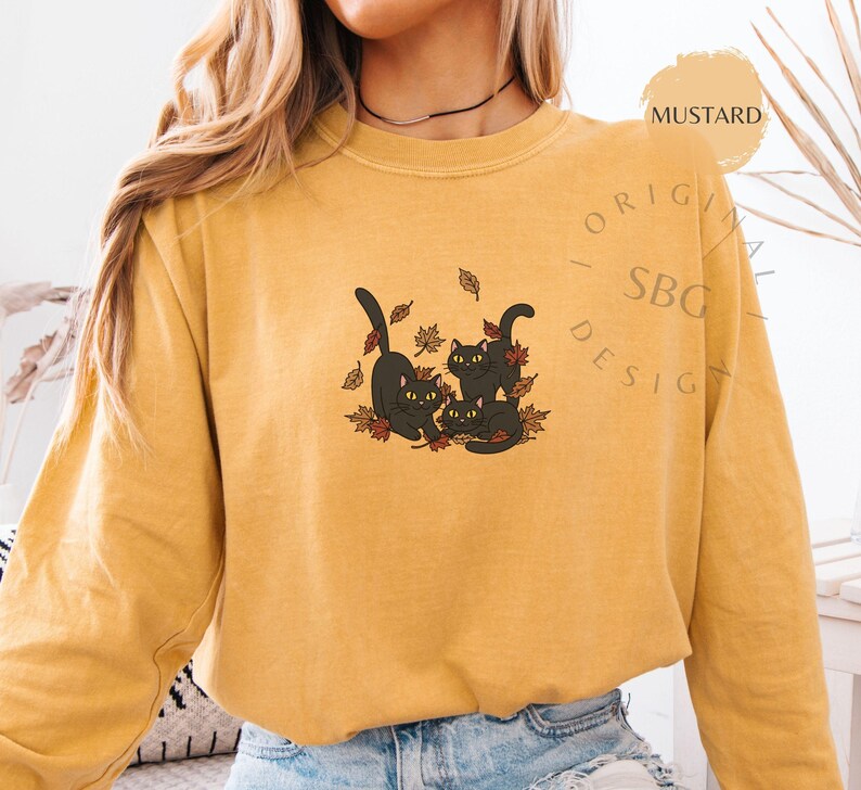Puede incluir: Sudadera de color mostaza con un gr&aacute;fico de tres gatos negros jugando entre hojas de oto&ntilde;o. El dise&ntilde;o incluye el texto "ORIGINAL SBG DESIGN". La sudadera est&aacute; hecha de un material suave.