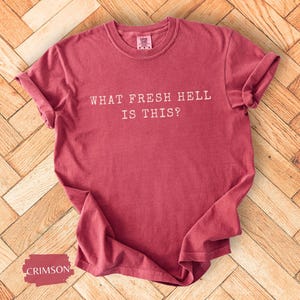 以下が含まれることがあります： 袖をまくったクリムゾン色のTシャツ。白い文字で「WHAT FRESH HELL IS THIS？」と書かれています。木製の表面に表示されています。