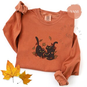 Puede incluir: Camiseta de manga larga naranja quemado con un gr&aacute;fico de tres gatos negros jugando entre hojas de oto&ntilde;o. Los gatos tienen ojos amarillos. La camiseta tiene cuello redondo y mangas enrolladas.