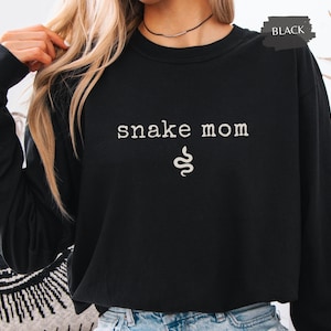May include: Black sweatshirt with the text "snake mom" in white, with a white snake graphic below the text. The word "BLACK" is in a black rectangle in the upper right corner. The sweatshirt is worn by a person.