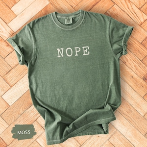 May include: Moss green t-shirt with the word "NOPE" in white block letters. The shirt has a crew neck and short sleeves. The shirt is made of a soft, comfortable material.