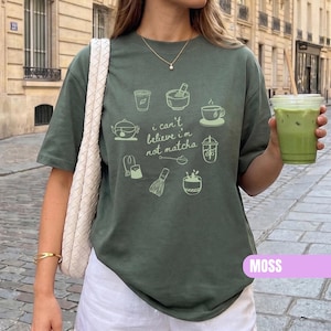 May include: A moss green t-shirt featuring a light green graphic print of various tea and matcha preparation tools, including a whisk, bowl, and cup, with the text "i can't believe i'm not matcha" centered among the illustrations.