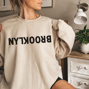 May include: A beige sweatshirt with the word "BROOKLYN" printed in black letters, reversed.