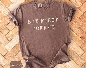 May include: Espresso-coloured t-shirt with the text "BUT FIRST COFFEE" in white block letters. The shirt is laid flat on a wooden surface. The shirt has short sleeves and a crew neck.