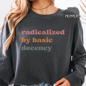 Könnte beinhalten: Ein anthrazitfarbenes Langarm-Sweatshirt mit dem Text "radicalized by basic decency" in drei untereinander gedruckten Zeilen. Das Wort "radicalized" ist rosa, "by basic" ist orange und "decency" ist salbeigrün. Ein rundes Etikett zeigt "PEPPER".