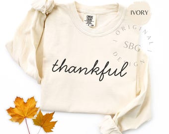 May include: Ivory long-sleeve shirt with the word "thankful" in black script. Two yellow and orange autumn leaves are placed near the bottom of the shirt. The shirt has a Comfort Colors tag.