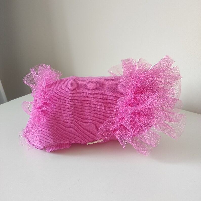 Peut inclure: Un lit pour animaux de compagnie rose vif, de forme rectangulaire et &agrave; texture c&ocirc;tel&eacute;e. Le lit est orn&eacute; de tissu rose &agrave; volants et transparent avec des pois blancs &agrave; chaque extr&eacute;mit&eacute;. Le lit est pos&eacute; sur une surface blanche.