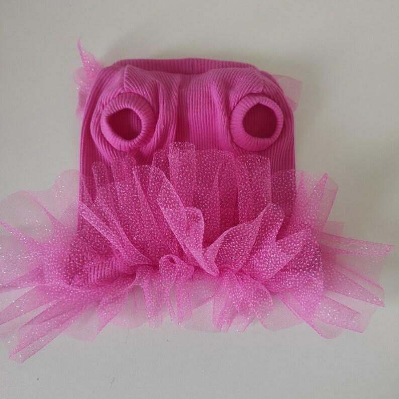 Peut inclure: Un pull rose vif pour chien, avec une texture c&ocirc;tel&eacute;e et des ouvertures pour les pattes avant. Il est orn&eacute; d'une jupe en tulle rose scintillante superpos&eacute;e, cr&eacute;ant un effet tutu. Le v&ecirc;tement est con&ccedil;u pour un petit chien.