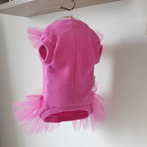 Peut inclure: Un pull pour chien rose vif avec une texture c&ocirc;tel&eacute;e, des manches courtes et une jupe en tulle. La jupe en tulle et les accents de manches sont d'un rose plus clair. Le pull est suspendu &agrave; un cintre en m&eacute;tal.