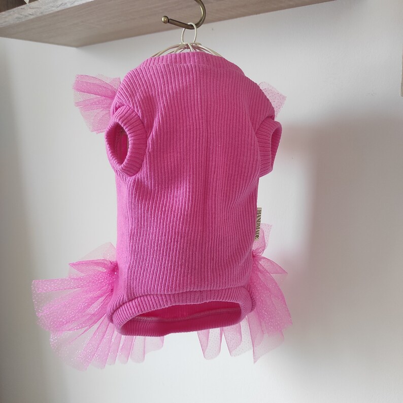 Peut inclure: Un pull pour chien rose vif avec une texture c&ocirc;tel&eacute;e, des manches courtes et une jupe en tulle. La jupe en tulle et les accents de manches sont d'un rose plus clair. Le pull est suspendu &agrave; un cintre en m&eacute;tal.