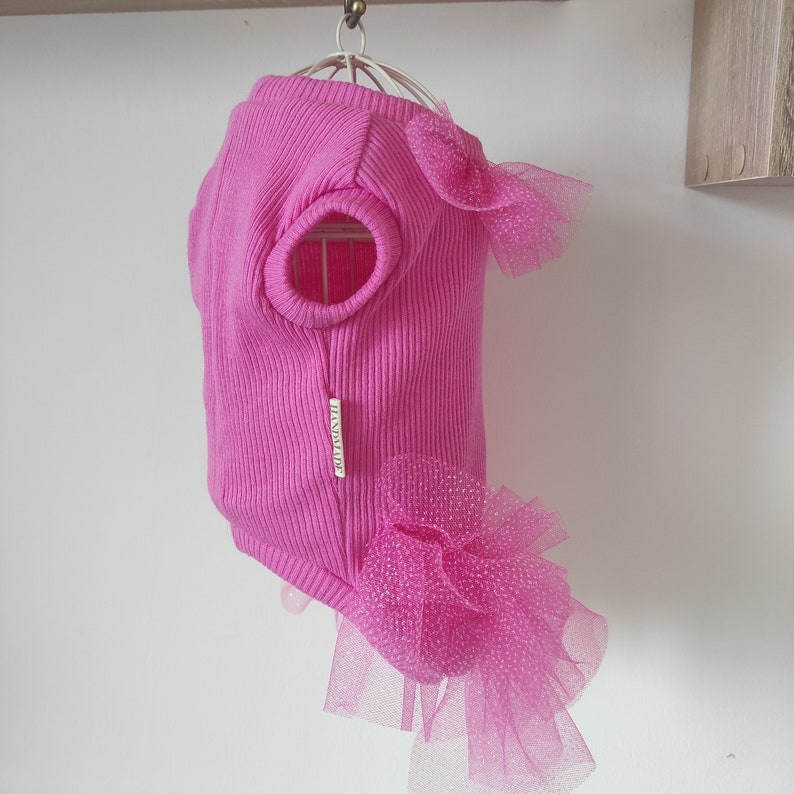 Peut inclure: Un pull pour chien rose vif &agrave; texture c&ocirc;tel&eacute;e. Le pull porte une petite &eacute;tiquette avec l'inscription "HANDMADE". Il est orn&eacute; de couches de tulle rose et d'un n&oelig;ud. Le v&ecirc;tement est con&ccedil;u avec des ouvertures pour les pattes du chien.