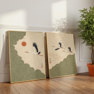 Könnte beinhalten: Zwei gerahmte Kunstdrucke mit japanisch inspirierten Illustrationen. Einer zeigt eine rote Sonne und einen Kranich, der über grünen Wolken fliegt. Der andere zeigt drei Kraniche im Flug über einer Landschaft mit grünen Hügeln. Beide haben goldene Rahmen.