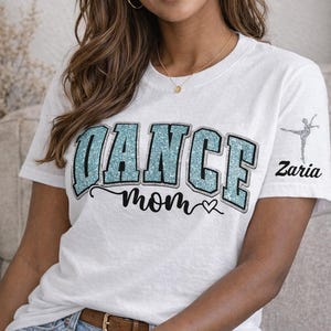 May include: A white t-shirt featuring the word DANCE in large, light blue glittery block letters with a black outline. Below, the word mom is written in black cursive script with a small heart. A dancer silhouette and the name Zaria appear on the sleeve.