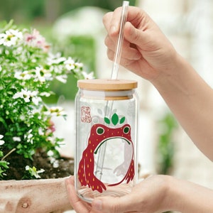 以下が含まれることがあります： 竹製の蓋と透明なストローが付いた透明なガラスタンブラー。タンブラーは、緑の葉と王冠が付いた赤いイチゴのカエルデザインが特徴です。タンブラーは人が持っています。
