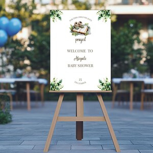 Op de afbeelding: Een houten ezel toont een welkomstbord voor een babyshower. Het bord bevat de tekst "Voor dit kind hebben we gebeden" en "Welkom bij Abigail's Babyshower" met de datum 25 december. Groen en babyschoentjes zijn afgebeeld.
