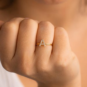 Könnte beinhalten: Ein goldfarbener Ring mit dem Buchstaben "A", besetzt mit kleinen, klaren Edelsteinen. Der Ring hat ein dünnes Band, und der Buchstabe steht im Mittelpunkt. Der Ring wird am Finger getragen und unterstreicht sein elegantes Design.