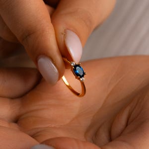 May include: A gold ring with a dark blue oval gemstone and two smaller white gemstones. The ring is held in a hand, highlighting its elegant design. The ring's design is delicate and the gemstones are set in a gold band.