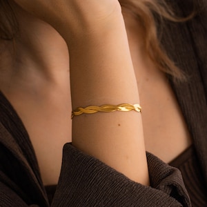 Könnte beinhalten: Ein goldfarbenes Armband mit geflochtenem Design. Das Armband befindet sich am Arm einer Person. Das Armband besteht aus einem glänzenden, metallischen Material. Das Armband ist ein Schmuckstück.