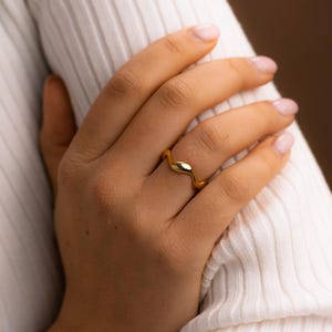 Könnte beinhalten: Ein goldfarbener Ring mit wellenförmigem Design. Der Ring befindet sich an einem Finger, vor einem weißen Pullover. Das Design des Rings weist eine längliche ovale Form in der Mitte auf.