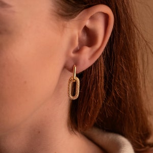 May include: Gold-toned oval drop earrings with a rectangular link design. The earrings feature a smaller, vertical bar connecting to a larger, embellished oval. The oval is encrusted with small, sparkling stones, creating a glamorous look. The earrings are shown on a person's ear.
